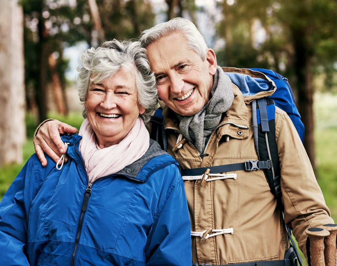 Smiling elderly couple outdoors wearing hiking jackets and backpacks.