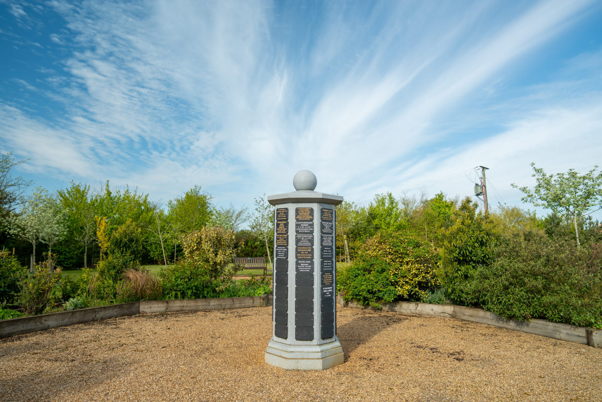 Granite memorial pillar with inscribed plaques on a gravel plot bordered by shrubs